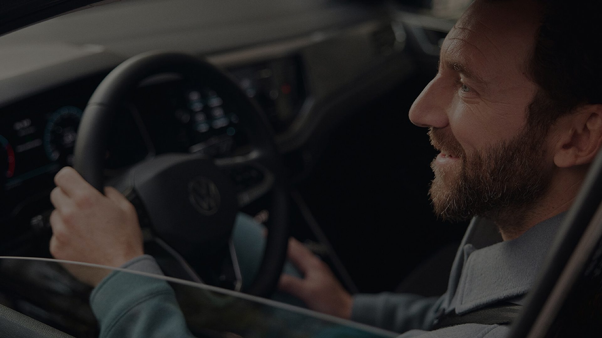 Un homme barbu et souriant, assis au volant, les mains sur le volant, regarde sur le côté.
