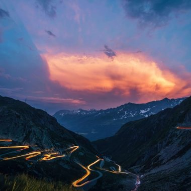 Route de montagne sinueuse au crépuscule, avec traces lumineuses de voitures et ciel dramatique orange et bleu.