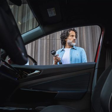 Un homme barbu tient une prise de charge devant un bâtiment, regardant sur le côté, vu depuis l’intérieur d’une voiture par la porte ouverte.