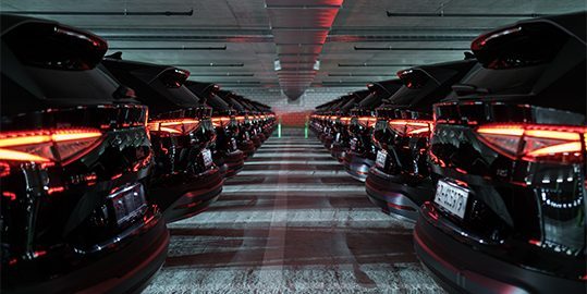 Une longue file de voitures noires identiques aux feux arrière rouges, garées dans un parking souterrain, s’étend au loin.
