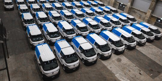 Vue en plan de nombreuses camionnettes blanches aux toits bleus, garées en rangées dans un grand hall.