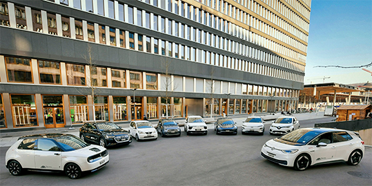 Plusieurs voitures électriques blanches sont garées sur une place devant un grand immeuble de bureaux moderne en ville.