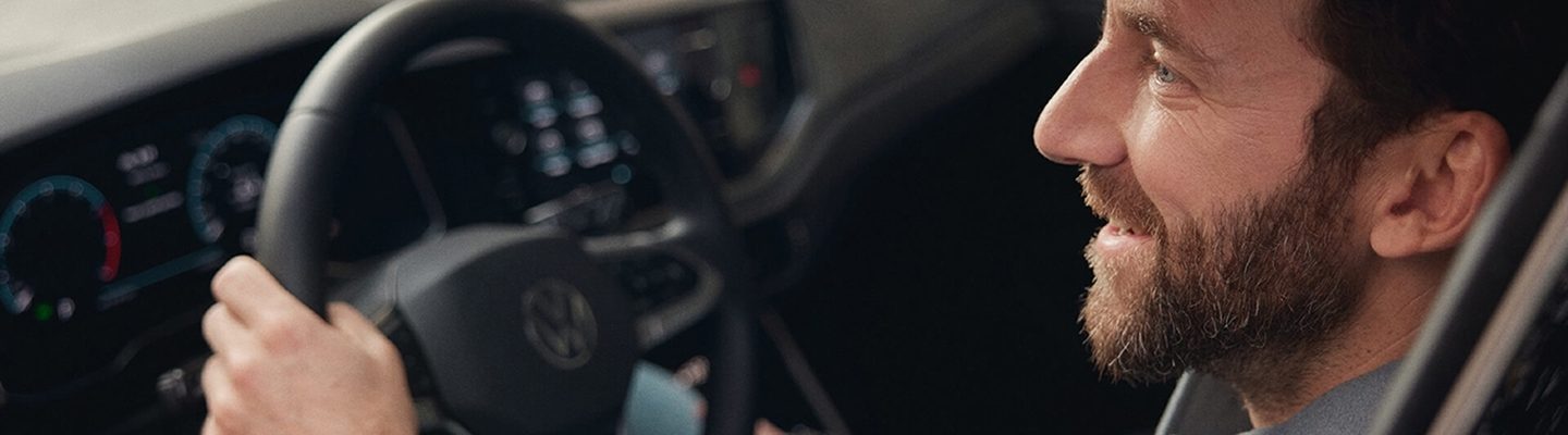 Ein lächelnder Mann mit Bart sitzt am Steuer eines Autos, seine Hände am Lenkrad, und blickt zur Seite.