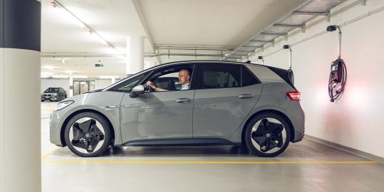 Ein Mann sitzt in einem grauen Elektroauto, das in einer Parkgarage neben einer leuchtenden Wandladestation geparkt ist.