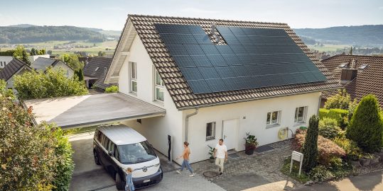 Ein modernes Haus mit Solarpaneelen auf dem Dach, davor ein Elektro-Van unter einem Carport und eine Familie im Vorgarten.