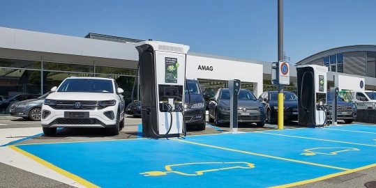 Eine Reihe von Elektroautos steht auf blau markierten Parkplätzen an mehreren weißen Schnellladestationen vor einem AMAG-Gebäude.