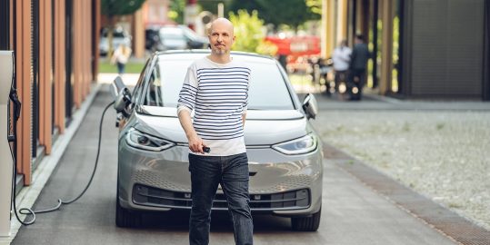 Ein Mann in einem gestreiften Shirt geht mit Autoschlüsseln auf den Betrachter zu, während im Hintergrund ein Elektroauto an einer Ladestation angeschlossen ist.