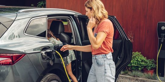 Eine Frau steht neben einem grauen Elektroauto, das lädt, während ein Kind im Fond des Wagens sitzt und zusieht.