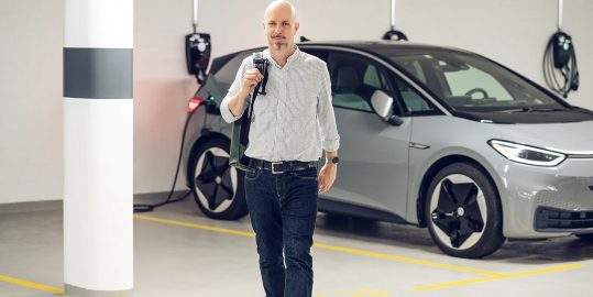 Ein Mann mit Tasche geht in einer Parkgarage auf den Betrachter zu, während im Hintergrund ein graues Elektroauto an einer Wandladestation lädt.